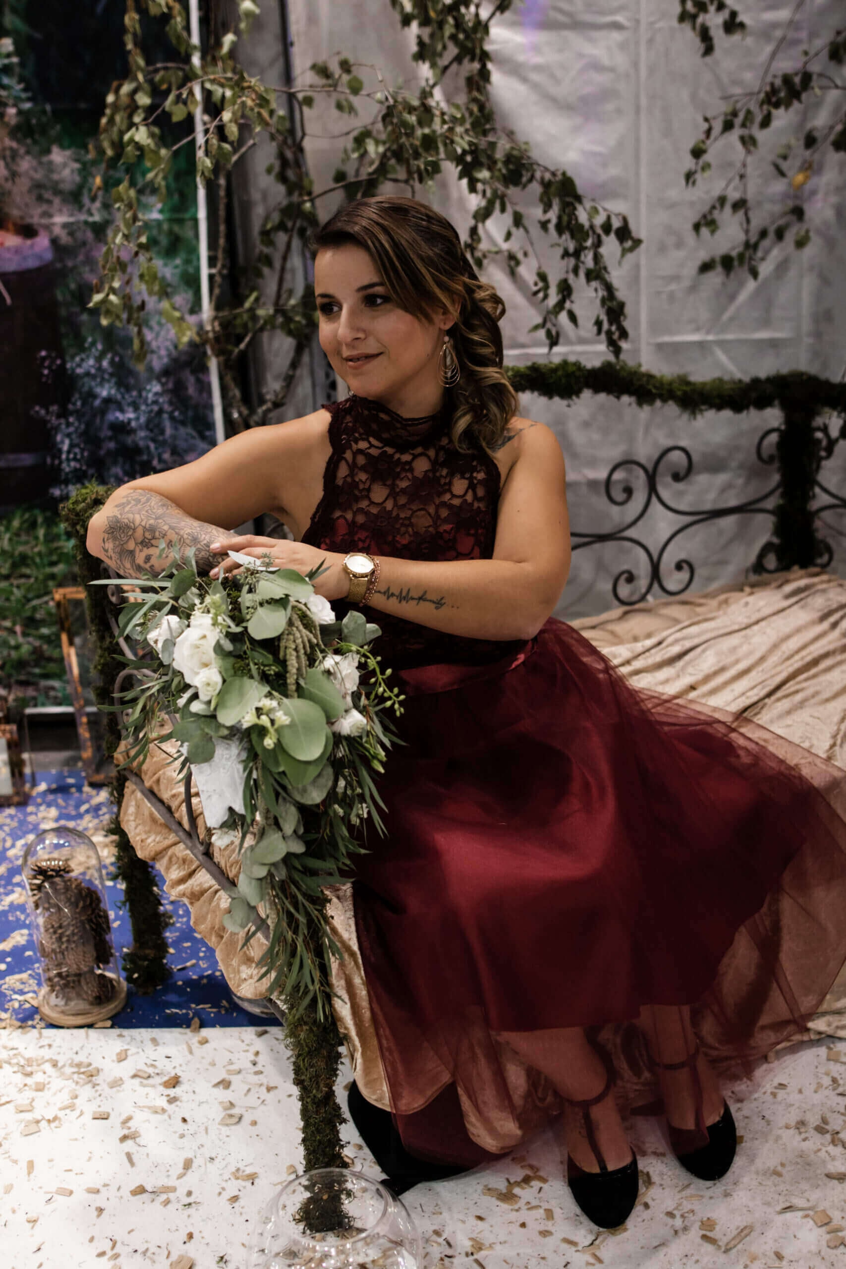 Une femme assise élégamment sur un banc décoré, portant un haut en dentelle noire sans manches et une jupe fluide bordeaux, entourée de verdure et d'un bouquet de fleurs blanches, dans une ambiance festive avec des décorations et des confettis éparpillés.
