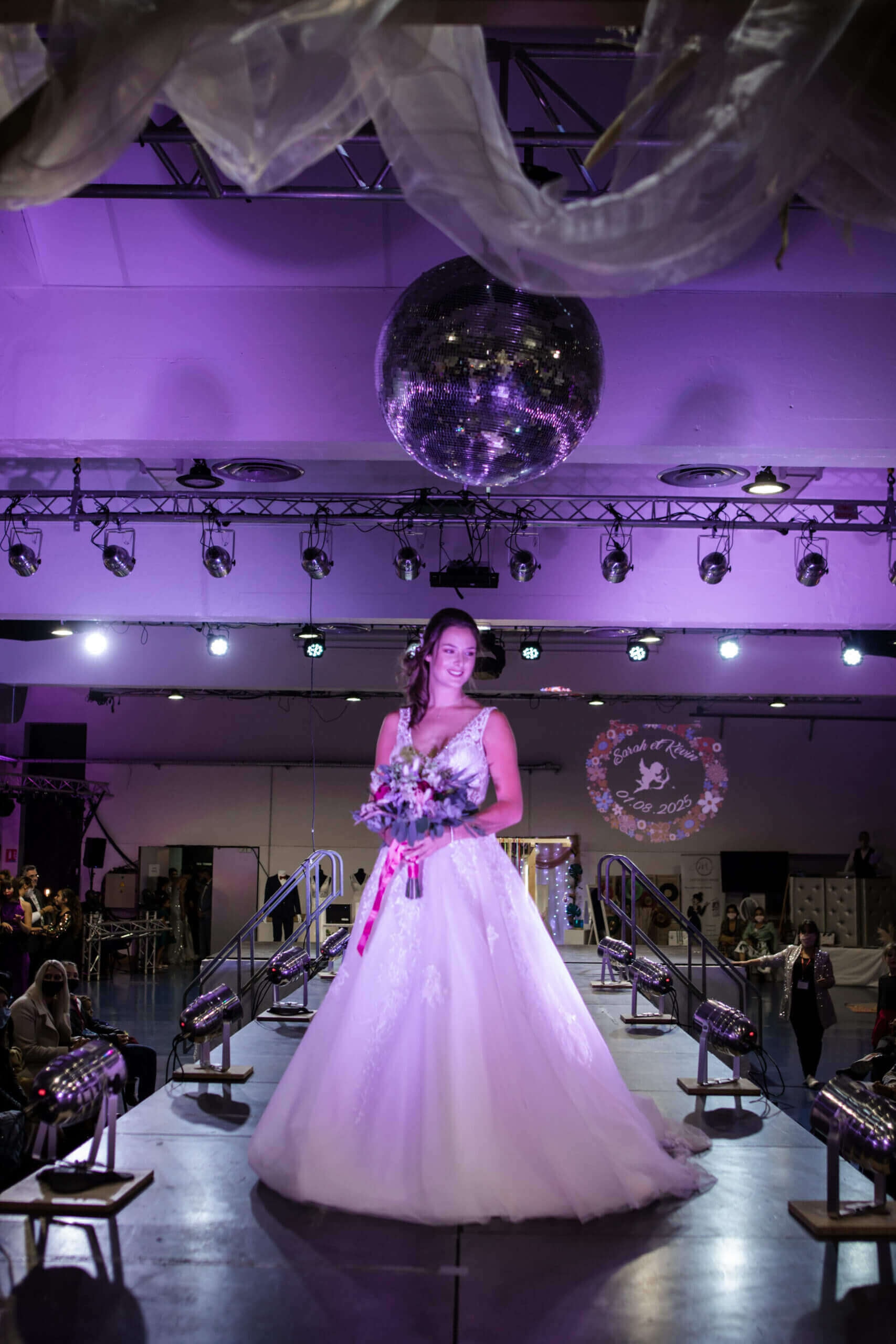 Une mariée se tient avec confiance sur le podium d'un défilé de mode, portant une élégante robe de mariée blanche ornée de détails complexes, tandis qu'elle tient un bouquet de fleurs et que ses cheveux sont coiffés en douces vagues, sous une lumière violette dramatique.