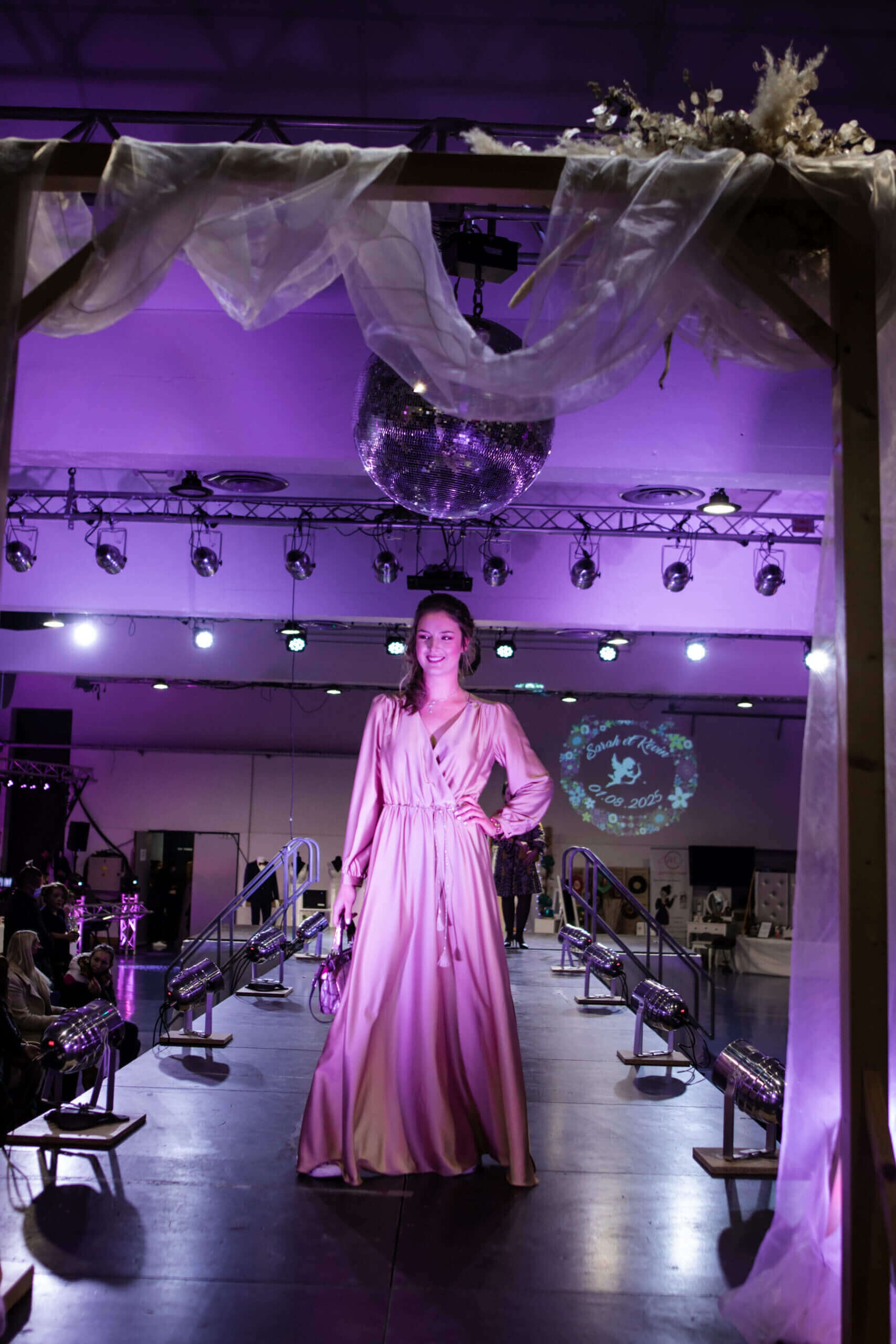 Un mannequin défile sur un podium de mode, portant une longue robe rose fluide sous une lumière violette, avec une boule disco suspendue et une arche décorative en tissu transparent, créant une ambiance glamour.