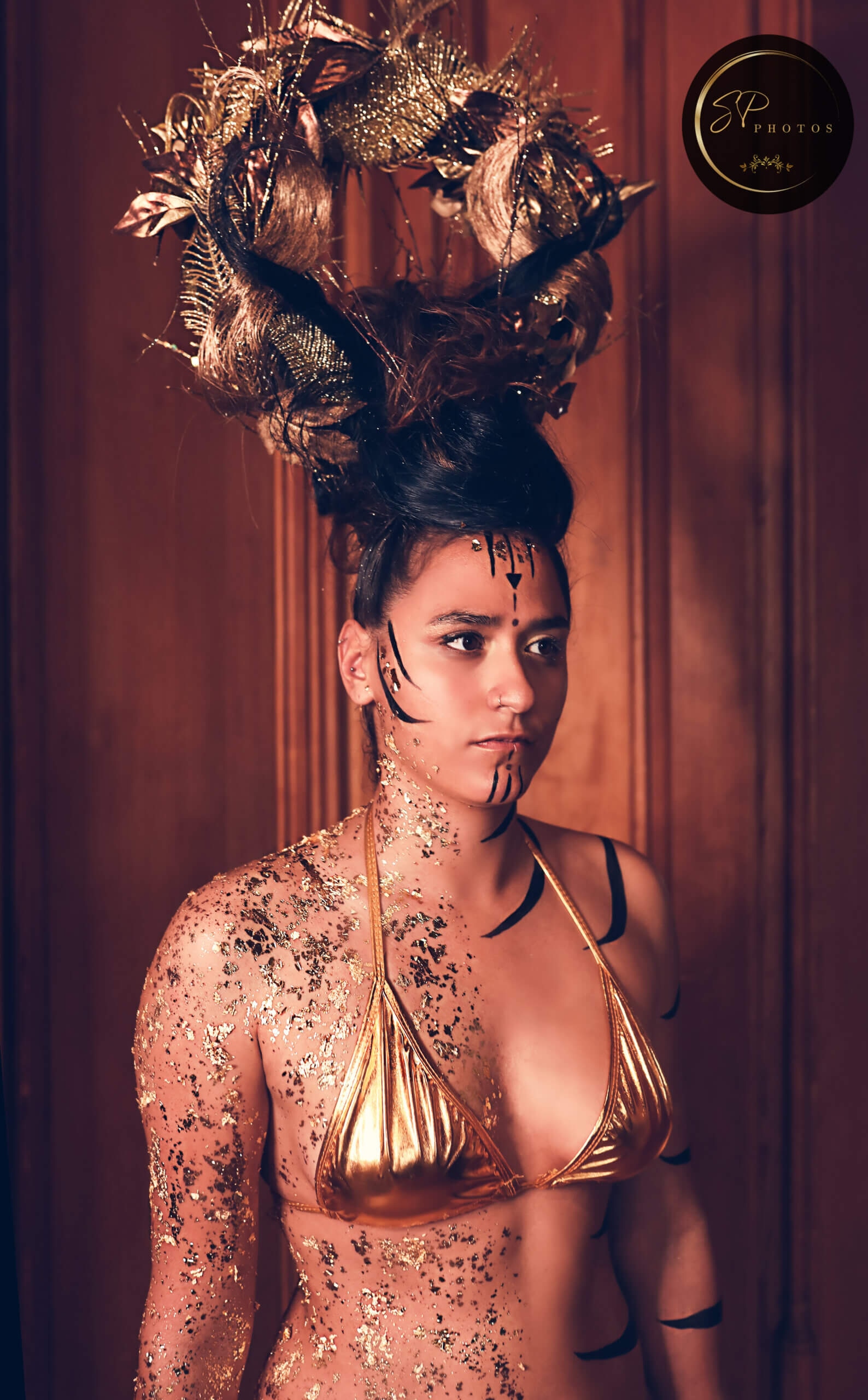 Une jeune personne se tenant devant un fond en bois, exhibant un look artistique saisissant avec un haut en bikini doré, de l'éclat doré sur la peau et une coiffure dramatique ornée d'un diadème élaboré en éléments dorés, ainsi que des marques artistiques audacieuses sur le visage, le tout baignant dans une lumière chaude qui souligne l'élégance du look.