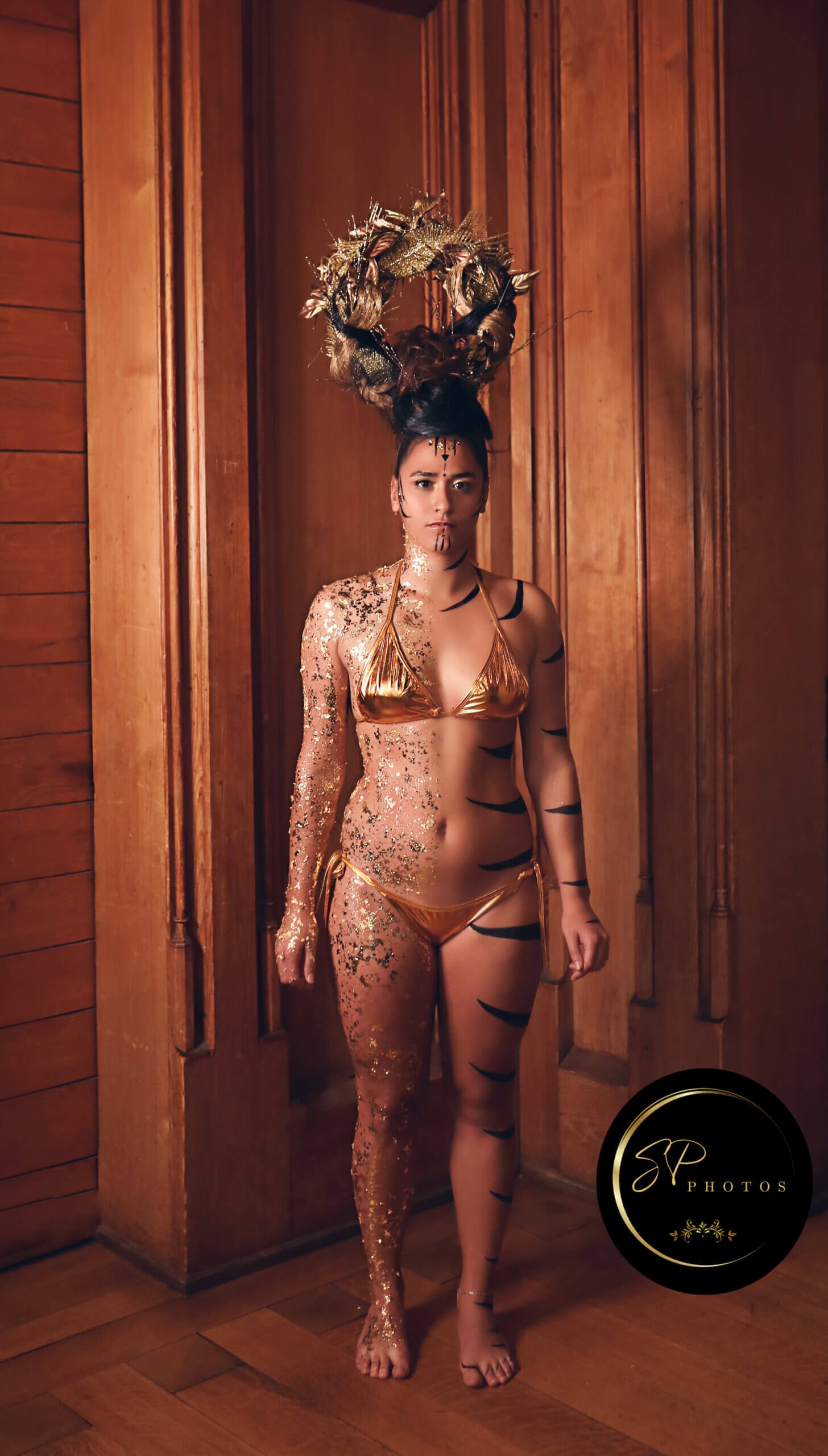 Un modèle se tient contre un fond en bois, arborant un bikini doré. Son corps est orné de peinture corporelle complexe en or et noir, ressemblant à des rayures de tigre. Elle porte un diadème frappant fait de feuillage et de brindilles, accentuant son allure audacieuse. L'éclairage met en valeur ses traits du visage et les textures de l'art corporel, tandis que le sol en bois crée une atmosphère chaleureuse. L'image véhicule des thèmes de créativité et d'expression artistique.