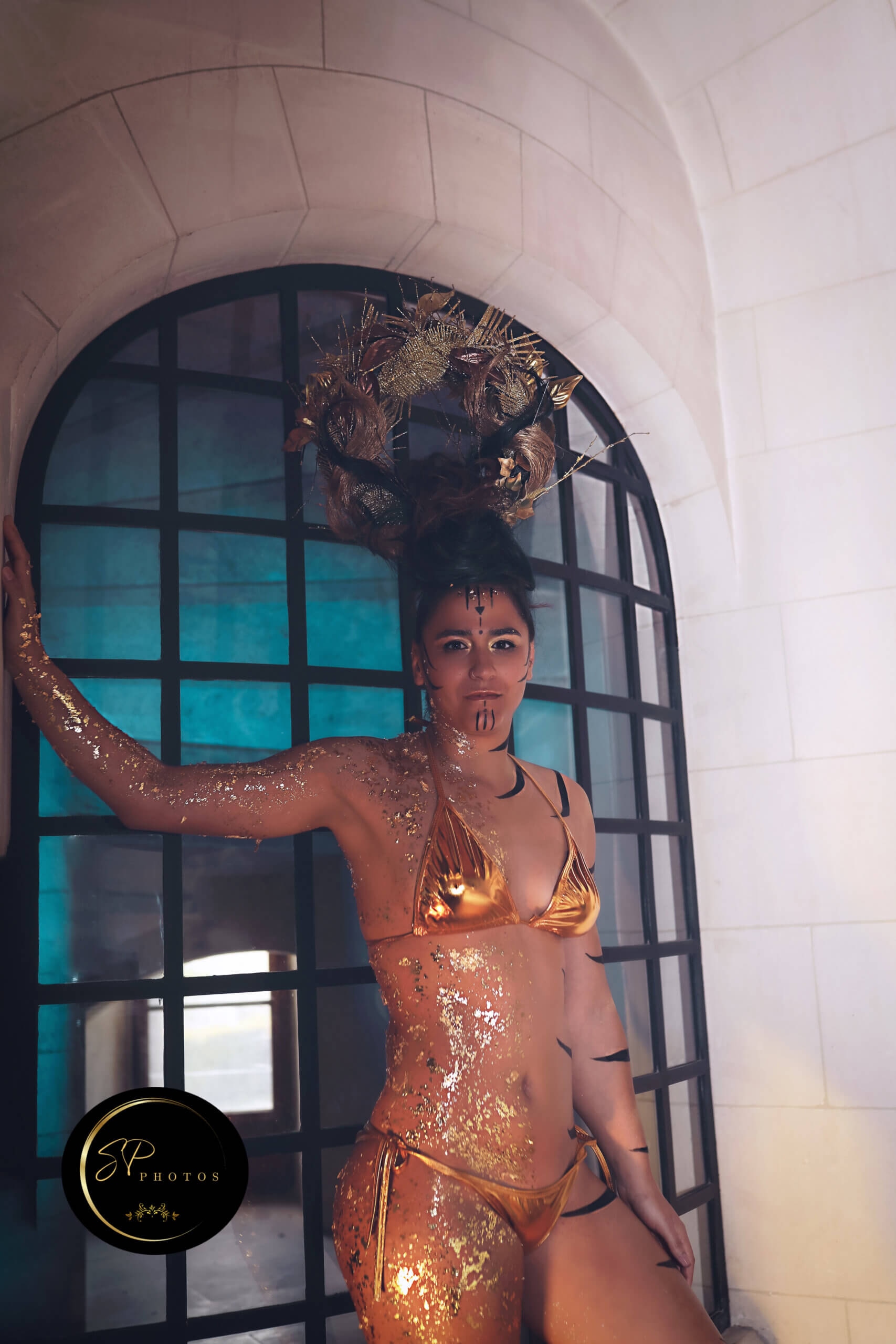 Une femme posant devant un mur en pierre dans une pièce semi-obscure, mettant en valeur son bikini doré scintillant et son corps orné de paillettes. Elle porte un maquillage tribal complexe et un superbe accessoire pour la tête avec des accents dorés, tandis qu'une grande fenêtre avec un éclairage bleu crée une ambiance éthérée.