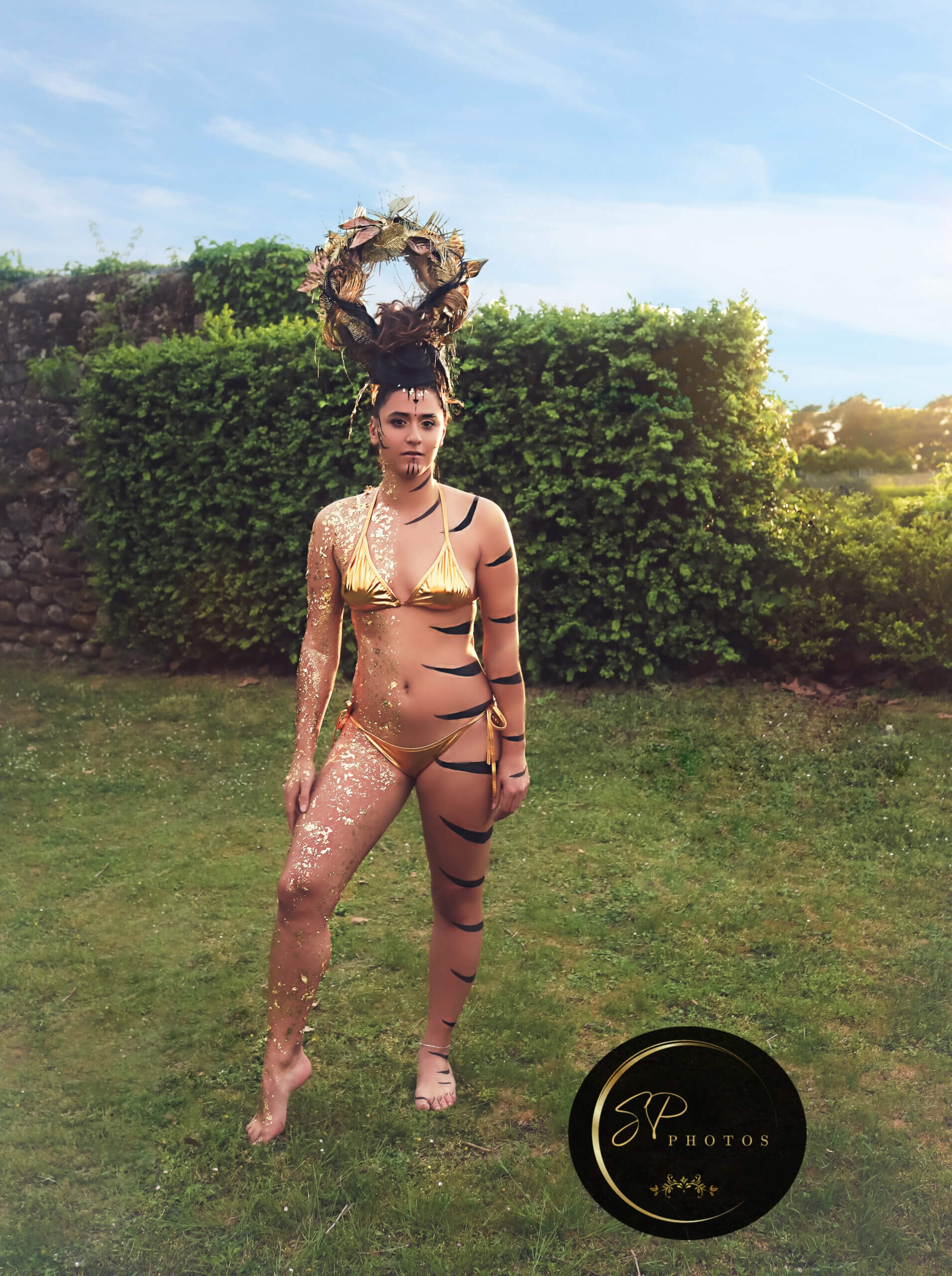 Une modèle confiante pose dans un cadre extérieur luxuriant, vêtue d'un bikini métallique doré, avec un maquillage corporel aux motifs animal insérés de paillettes dorées. Sa coiffure en chignon dramatique est ornée d'une couronne en feuilles, tandis que l'arrière-plan présente des haies taillées et un ciel bleu clair, créant une atmosphère vibrante axée sur la nature et l'expression artistique.