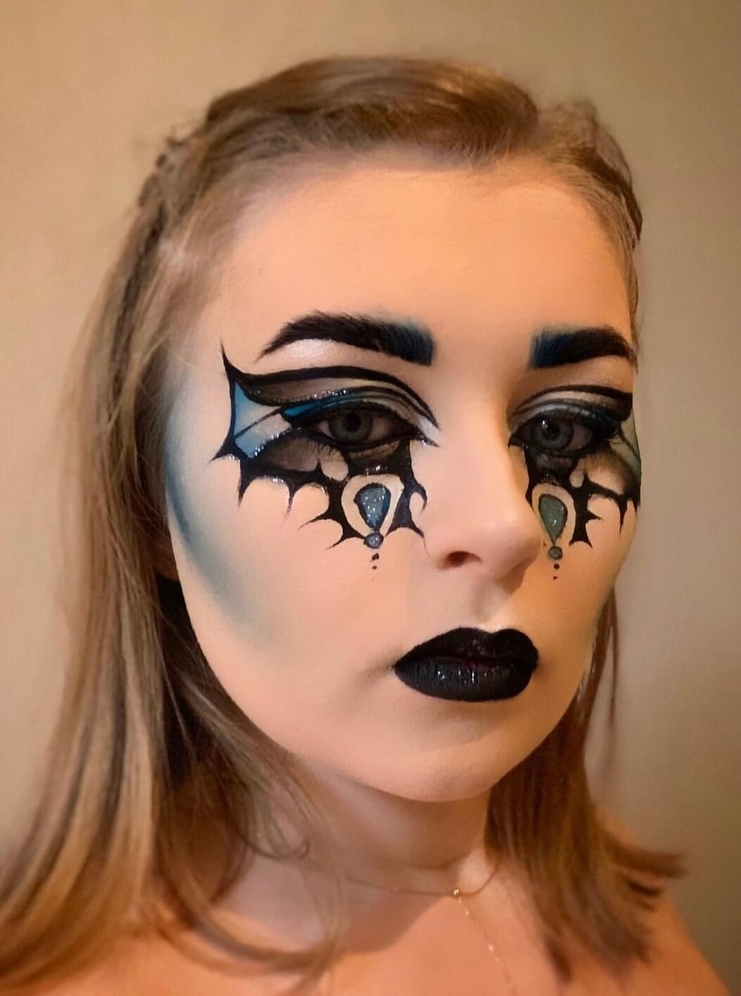 Une personne avec un maquillage artistique, présentant des motifs noirs et bleus qui ressemblent à des toiles d'araignées autour des yeux, accentués par un rouge à lèvres noir audacieux et des reflets bleus, le tout mis en valeur par une coiffure douce et décontractée.