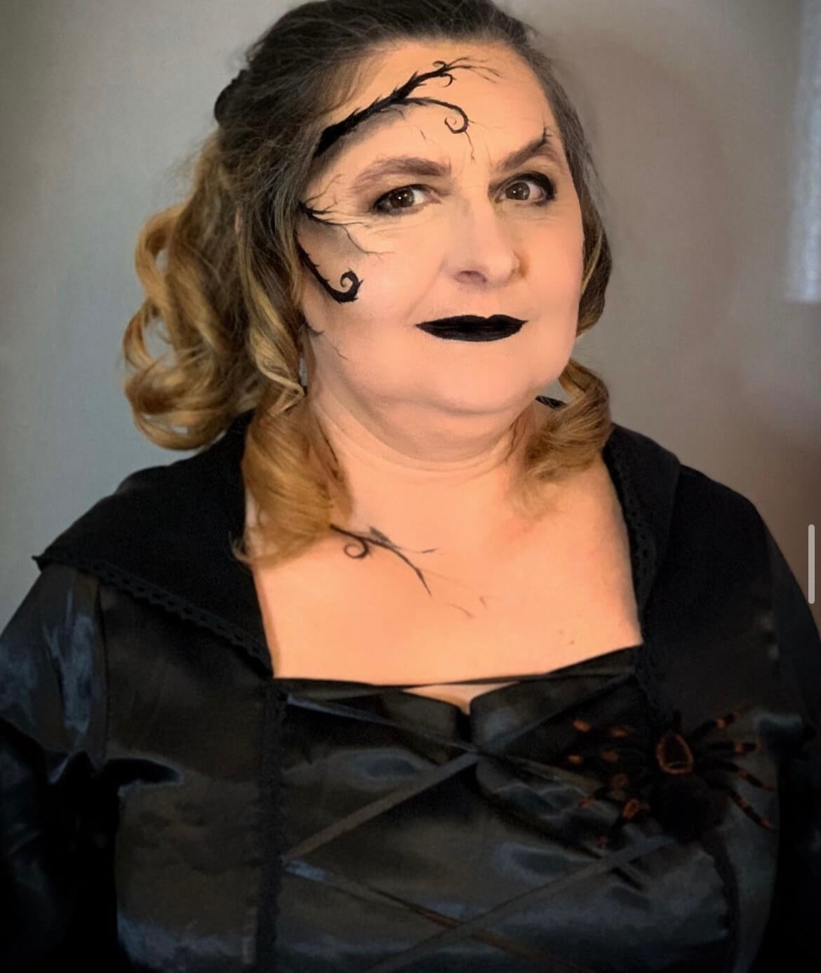 Une femme portant une robe noire élégante avec des détails décoratifs, ses cheveux en boucles douces avec des mèches grises, et un maquillage dramatique incluant du rouge à lèvres noir et des motifs de vignes sur son visage, posant avec confiance contre un fond neutre.