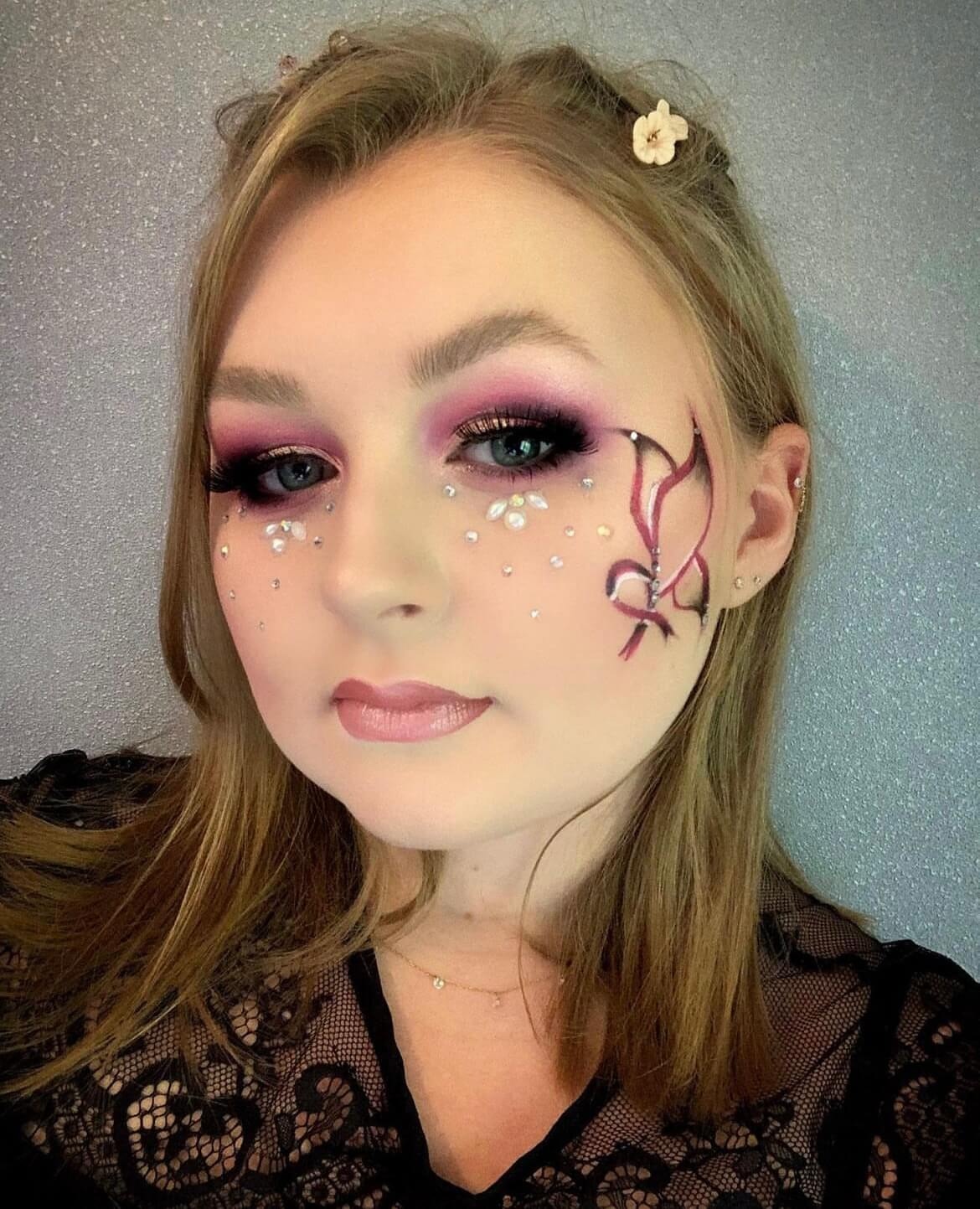 Une jeune femme avec un maquillage artistique et doux, affichant un smoky eye rose, des cils audacieux et des lèvres brillantes. Ses cheveux longs et châtains sont décorés de petites fleurs, tandis qu'un art facial complexe en forme de papillon est peint sur un côté de son visage, rehaussé de strass scintillants. Elle porte un haut en dentelle noire sur un fond argenté brillant.