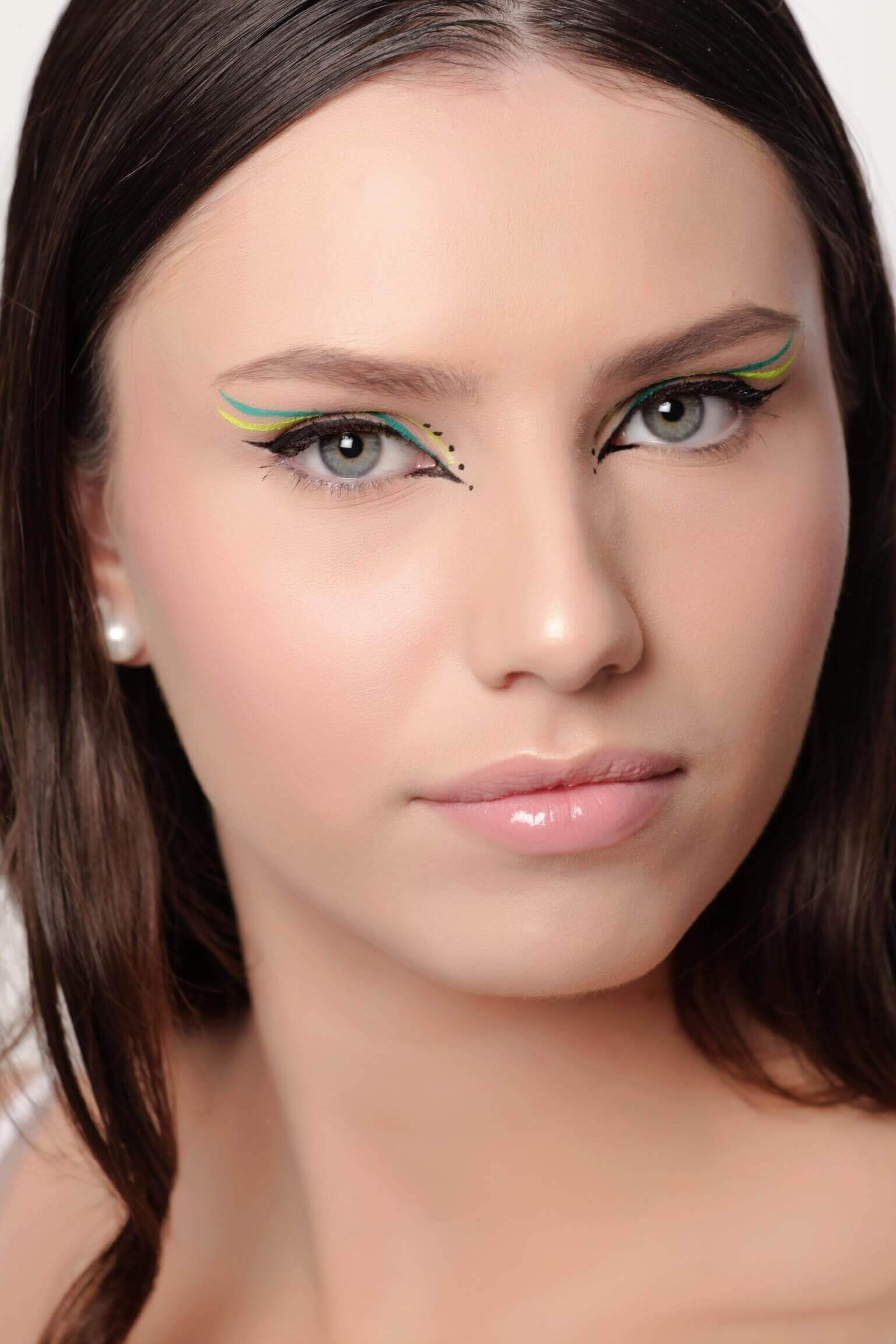 Une jeune femme avec de longs cheveux noirs et un maquillage neutre, mettant en avant un eye-liner vert et noir audacieux avec des motifs en pointillés sous les yeux, des lèvres glossées légèrement teintées, le tout sur un fond uni.