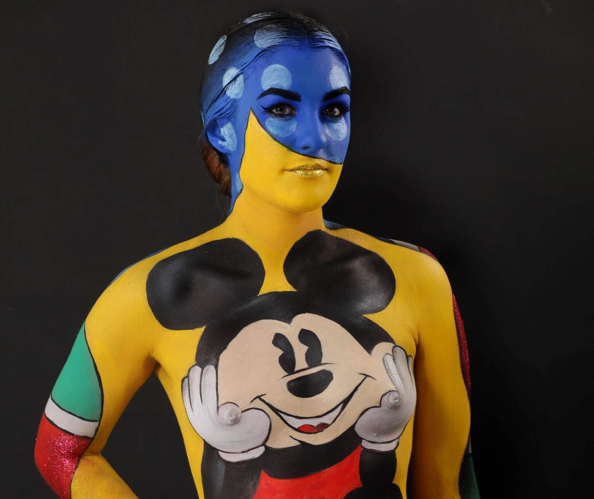 Un modèle pose devant un fond noir avec un design de peinture corporelle élaboré, mettant en avant un personnage de dessin animé coloré sur son torse, tandis que ses bras sont ornés de motifs multicolores et son visage est peint en bleu avec des accents dorés.