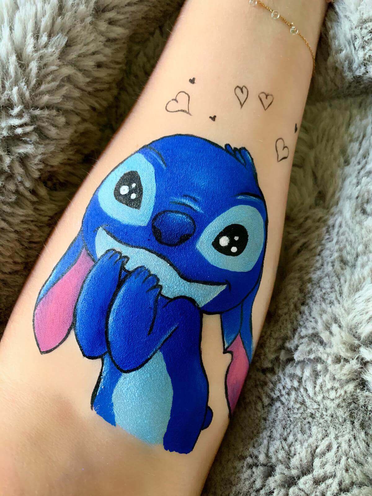 Un tatouage coloré et ludique sur un avant-bras, représentant un personnage de dessin animé bleu avec de grands yeux expressifs et une expression joyeuse, entouré de cœurs doodles dans une ambiance chaleureuse.
