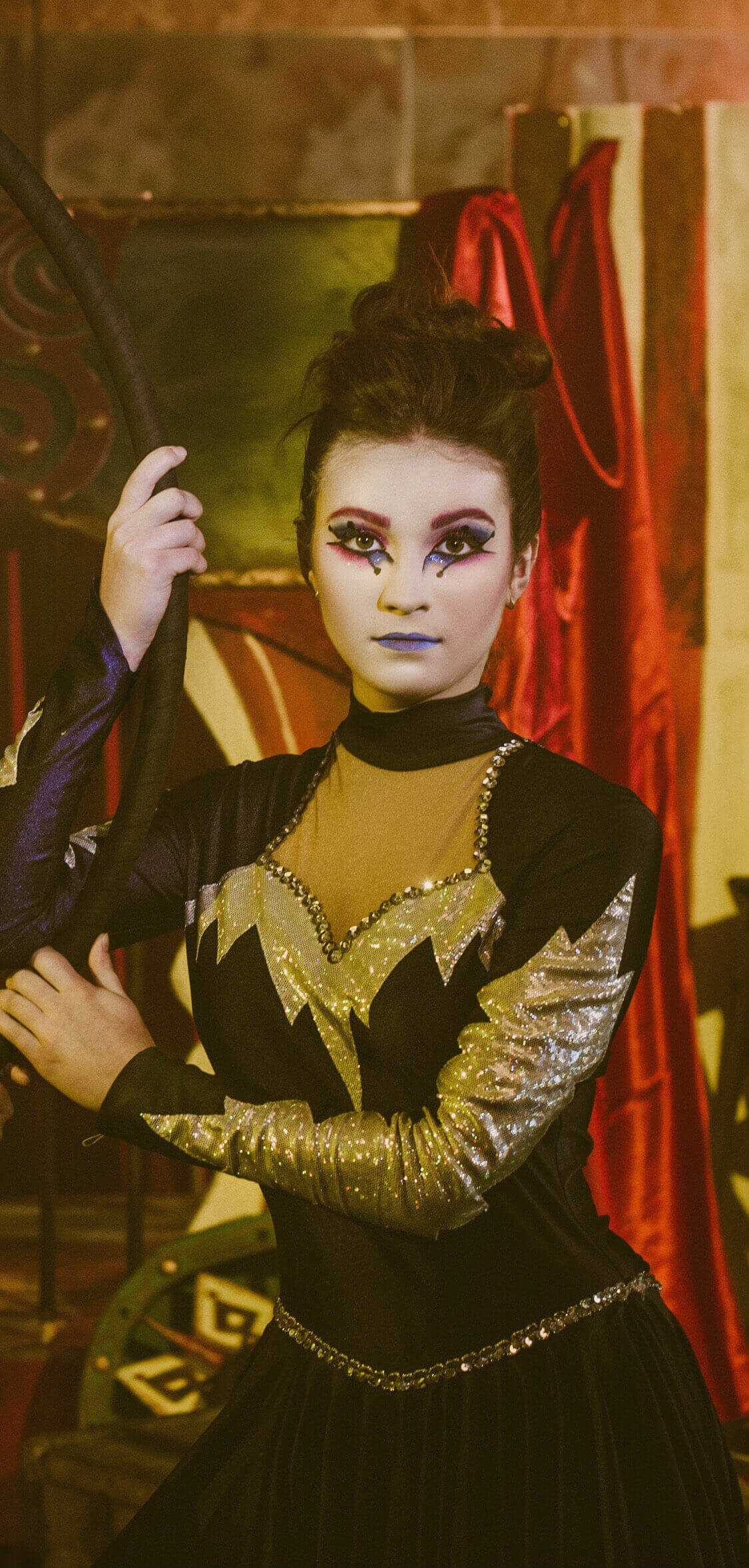Un artiste sur scène portant un costume noir avec des accents argentés, un maquillage audacieux et un fond théâtral avec des drapés rouges.
