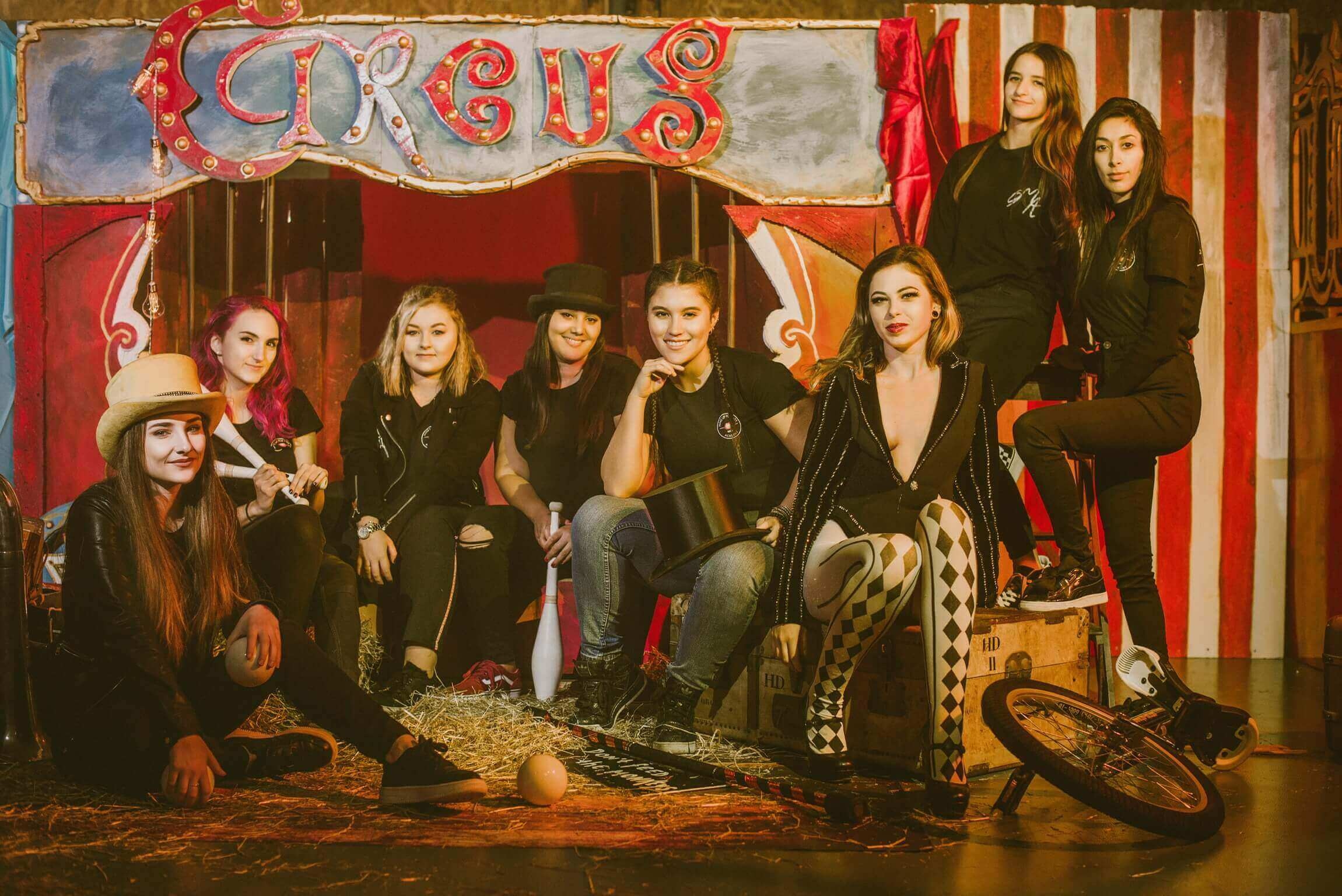 Un groupe de huit femmes s'amuse devant un fond coloré de cirque, avec des éléments en rouge et blanc, des accessoires de jonglage et une ambiance festive.