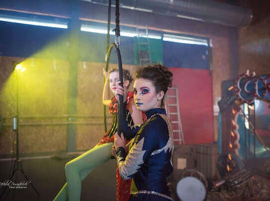 Deux artistes aériens en costumes colorés se produisent sur scène dans un décor de cirque, l'un suspendu gracieusement à une balançoire et l'autre debout avec assurance, dans une atmosphère vibrante accentuée par un éclairage doux et des accessoires de cirque.