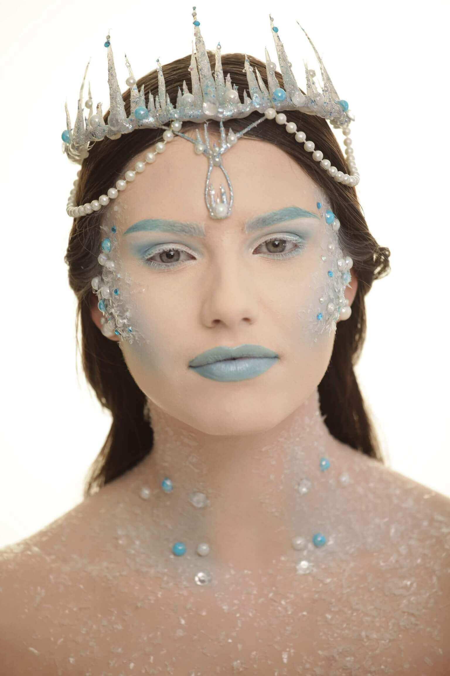 Portrait en gros plan d'une personne avec un maquillage et des accessoires élaborés sur le thème de la glace, présentant une peau pâle ornée de particules scintillantes et de gemmes bleues, avec des lèvres brillantes de couleur bleu clair et des yeux accentués par un fard à paupières bleu clair, le tout surmonté d'une couronne de perles et de formes cristallines, dans un arrière-plan illuminé et rêveur.