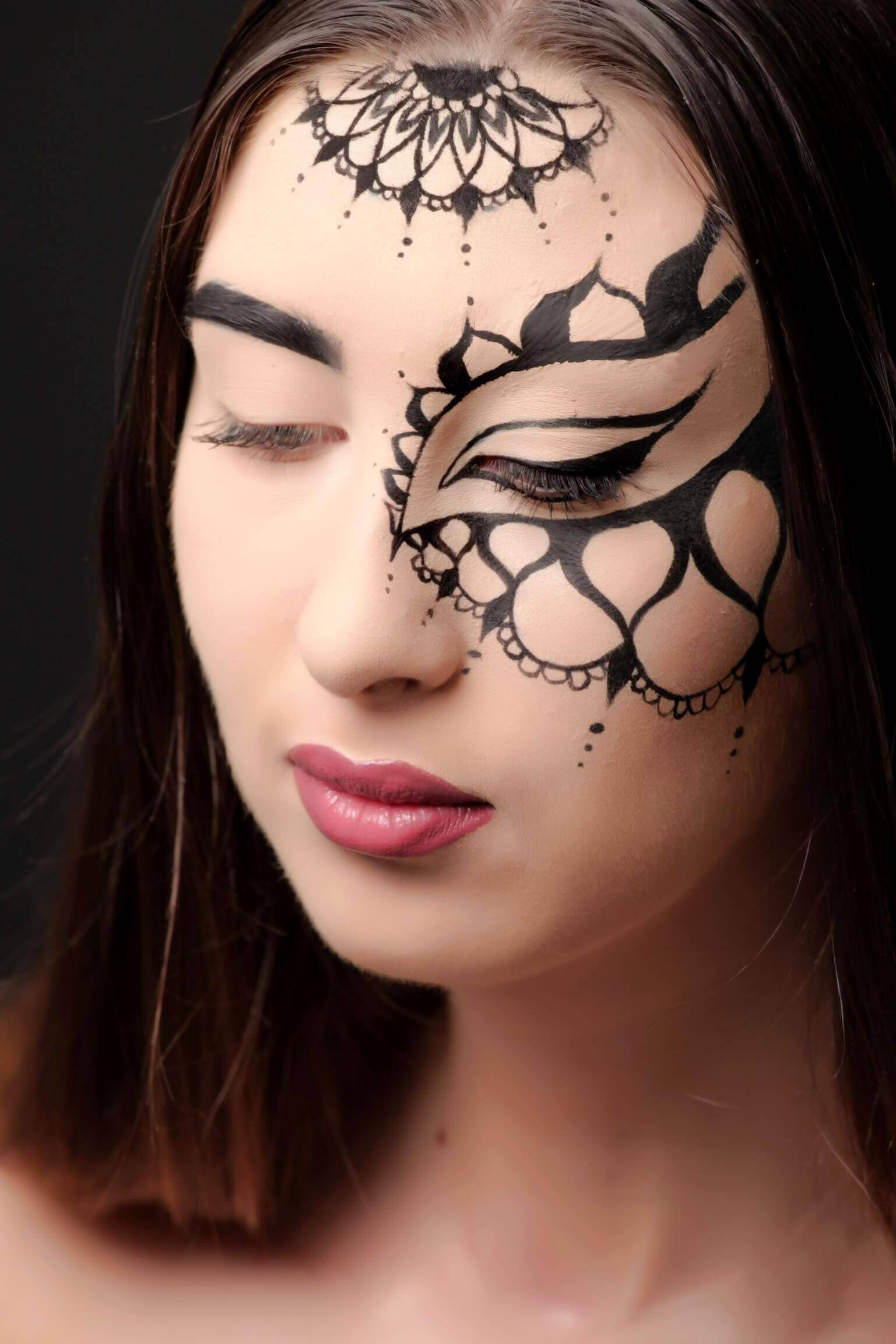 Un portrait en gros plan d'une femme avec des motifs artistiques noirs couvrant son front et sa joue, comprenant un œil stylisé et des éléments décoratifs inspirés de la dentelle et des motifs floraux, sur fond sombre.