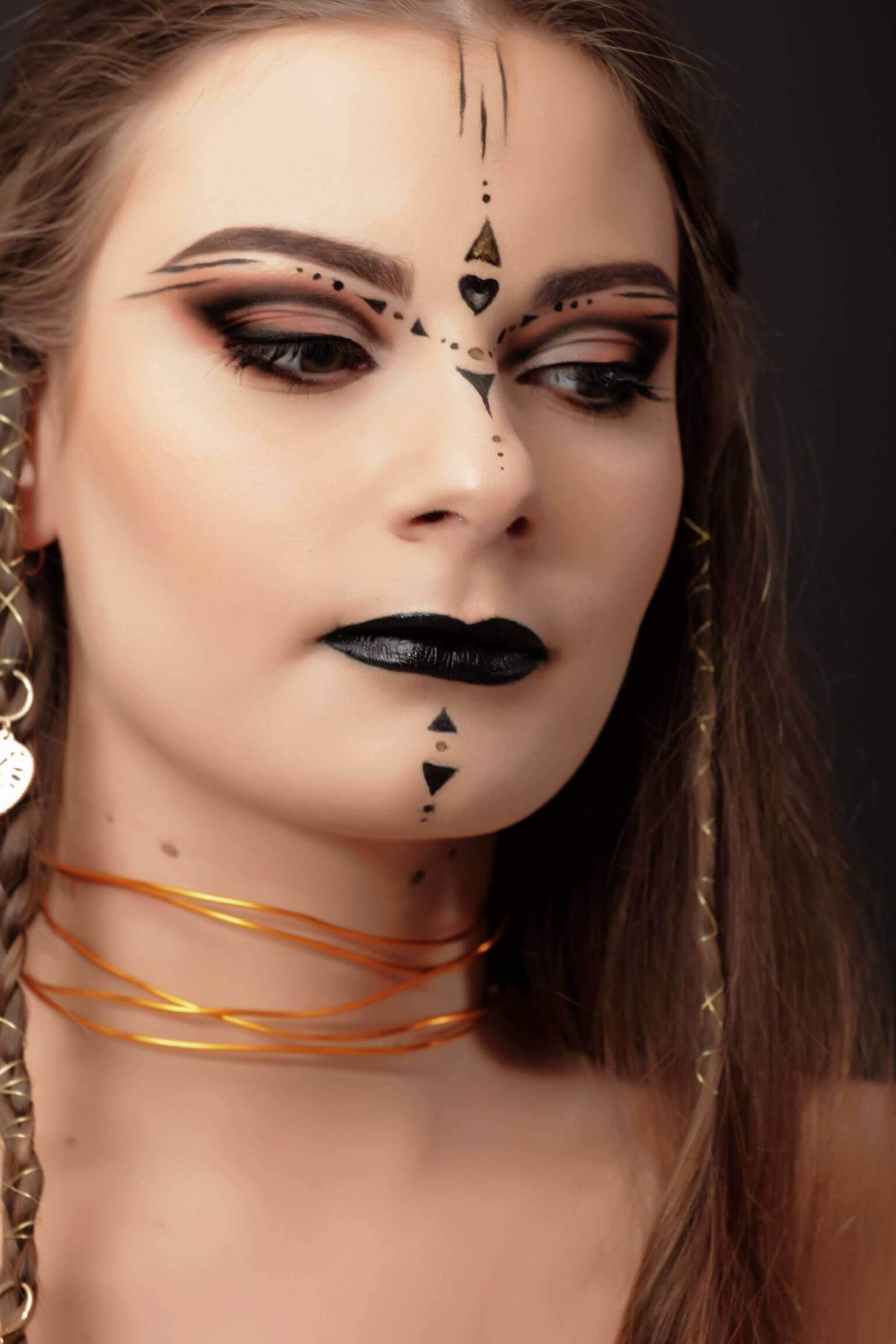 Un portrait en gros plan d'une femme avec un maquillage dramatique et des accessoires uniques, mettant en valeur des sourcils accentués et des designs d'eyeliner complexes, entourés d'une ombre brune chaude, des lèvres noires profondes, et des motifs géométriques frappants en noir et or sur son front et son menton.