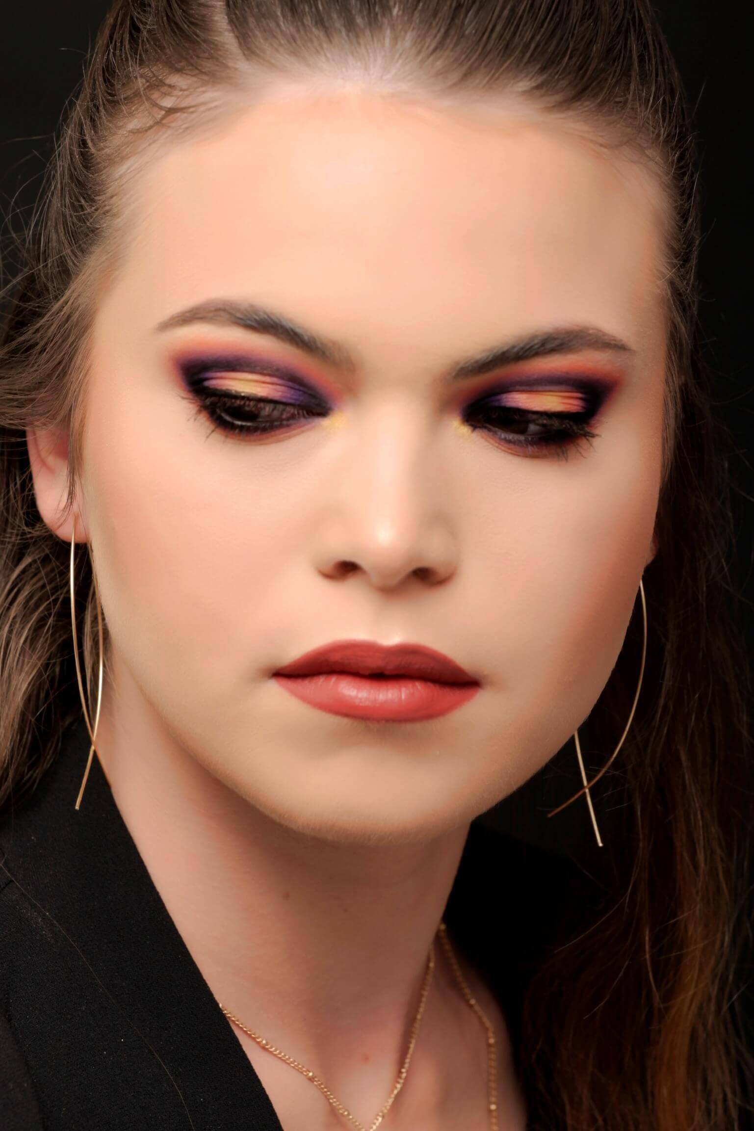 Portrait en gros plan d'une femme avec un maquillage des yeux dramatique aux teintes violettes et oranges, un teint lisse, des lèvres nude et des boucles d'oreilles en or, le tout sur fond sombre.