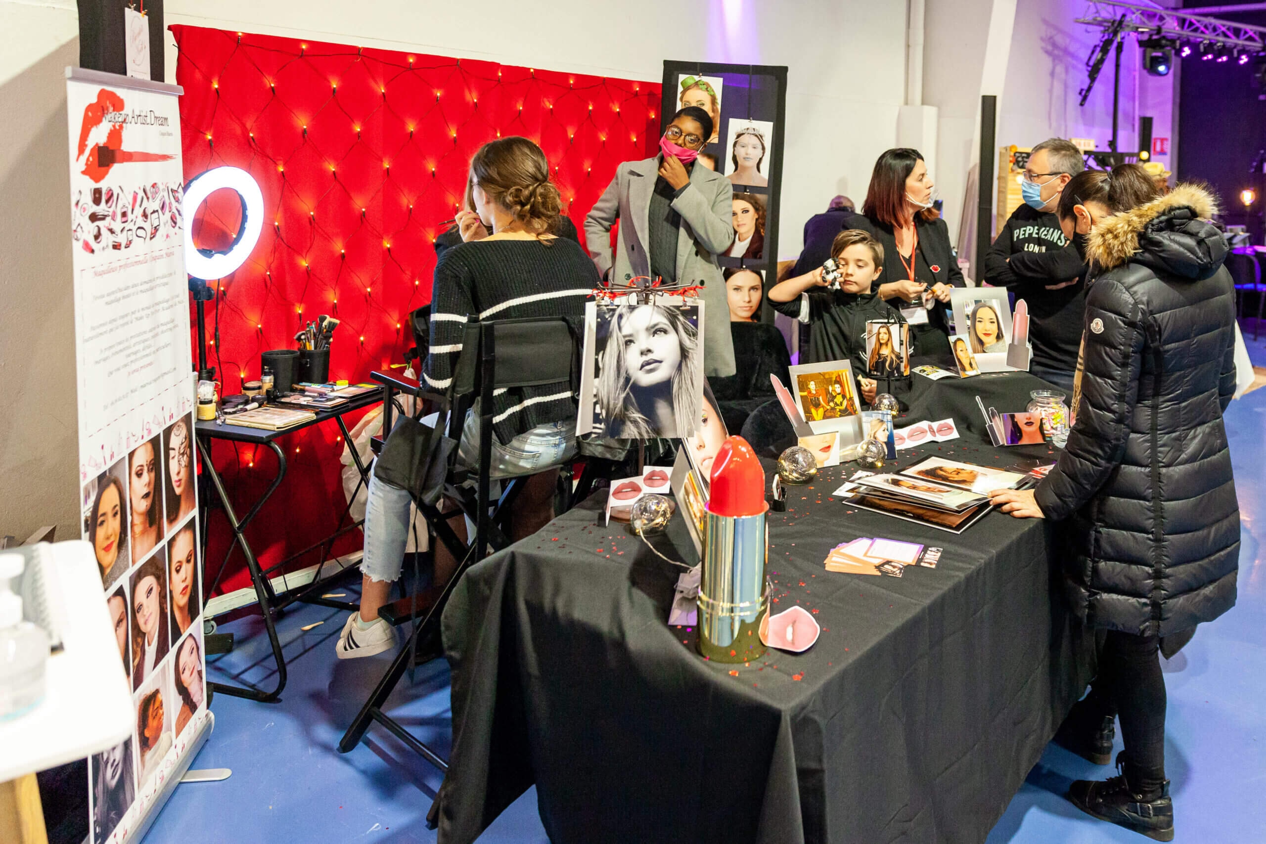 Un stand de maquillage vibrant lors d'un événement, avec une table remplie de produits de maquillage et des photos de différents looks, où une personne est en train de se faire maquiller et d'autres, y compris des adultes et un enfant, discutent et observent dans une ambiance vivante et interactive, soulignant la beauté et l'art.