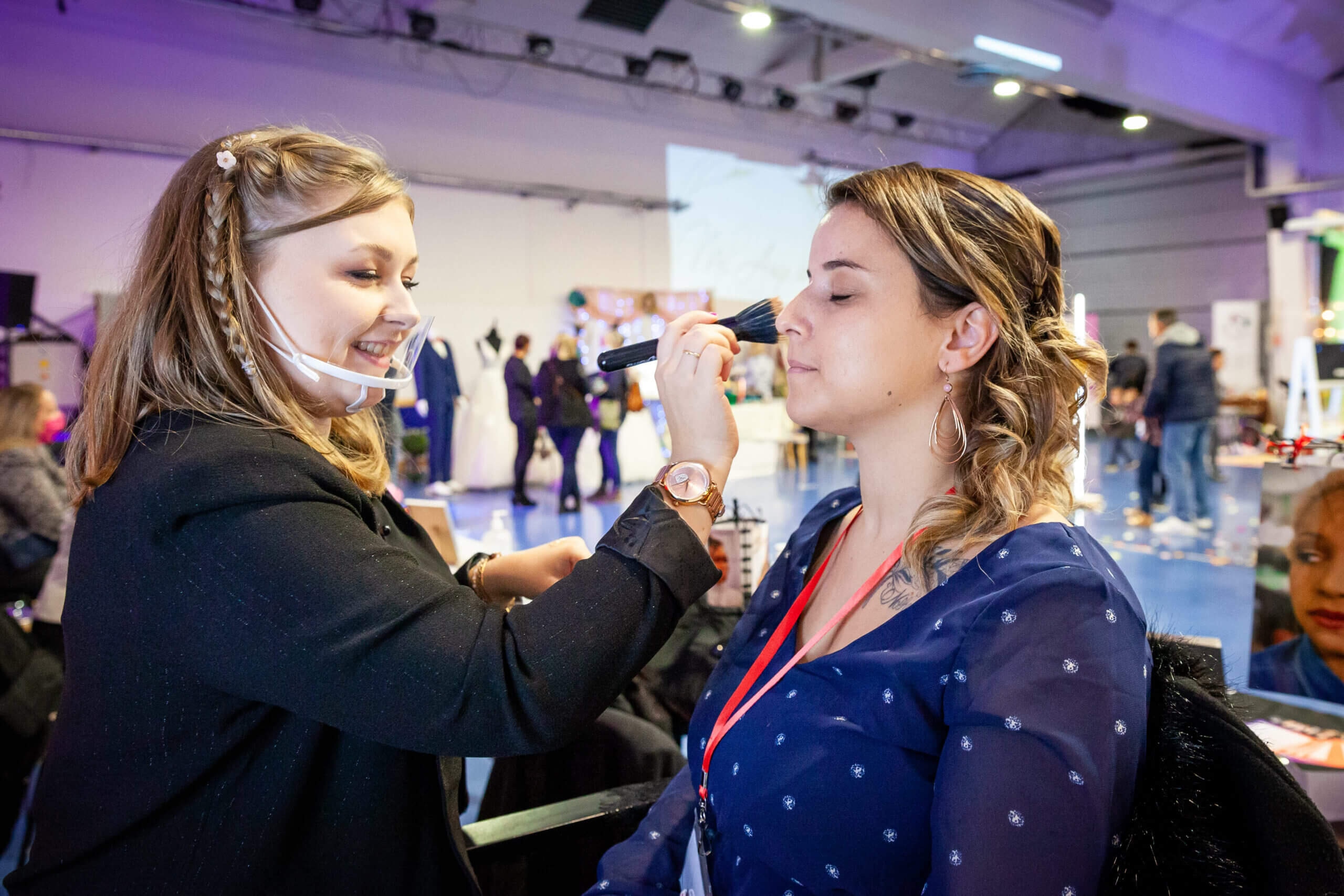 Une professionnelle de la beauté applique du maquillage à une cliente dans un cadre d'événement animé, avec des décorations colorées en arrière-plan.
