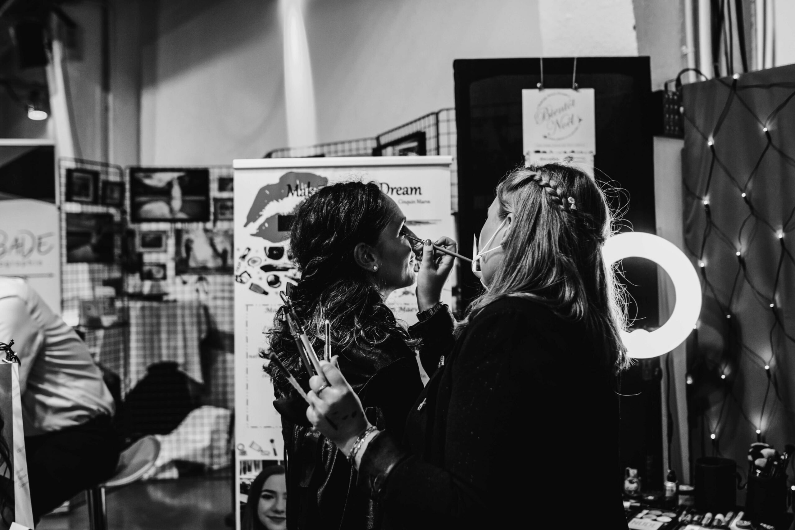 Deux femmes appliquent du maquillage dans un cadre intérieur vibrant, mettant en avant leur interaction intime sous une lumière circulaire, tandis qu'un fond artistique coloré les entoure.