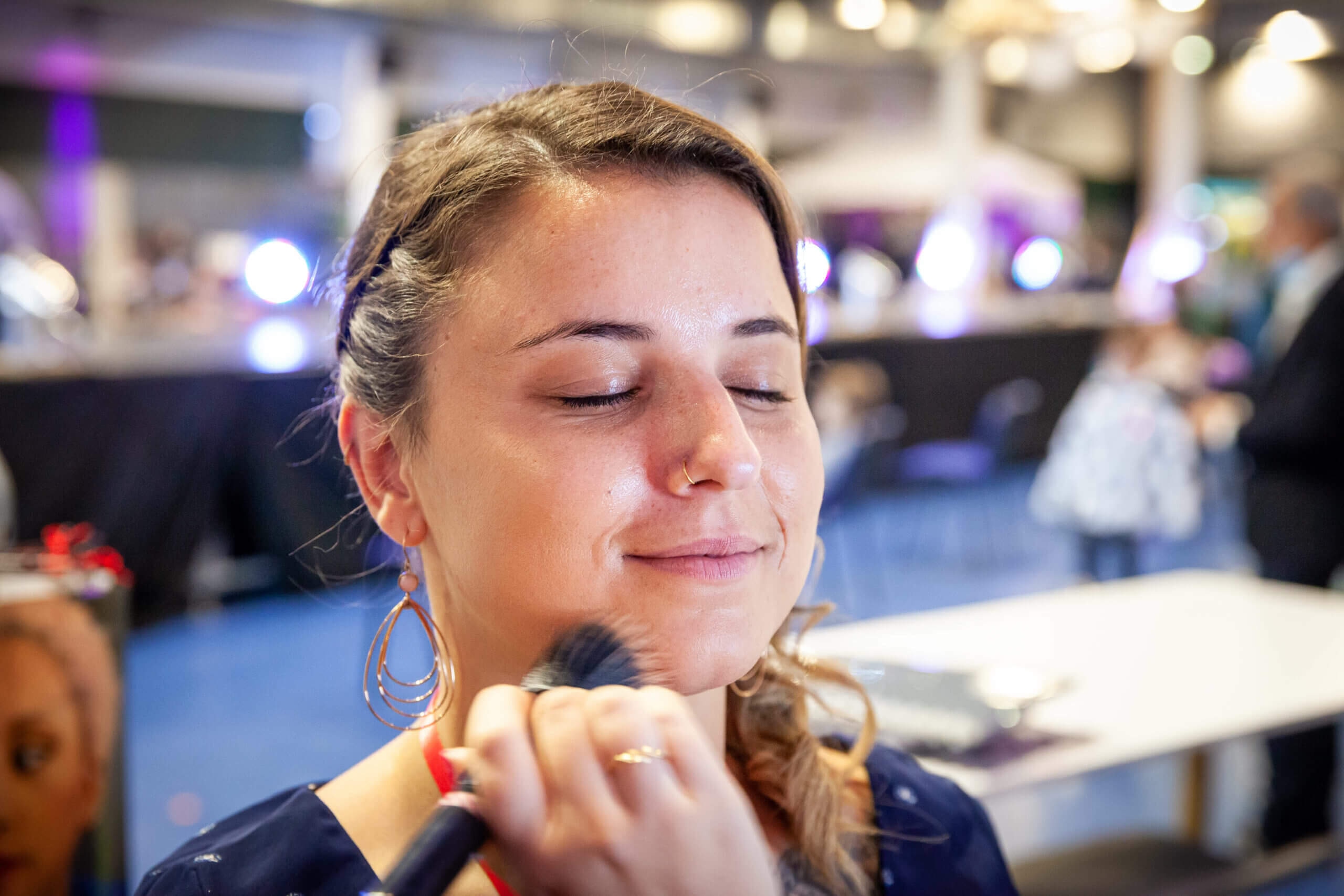 Une femme aux longs cheveux ondulés et portant des boucles d'oreilles en forme de hoop ferme les yeux avec un sourire satisfait pendant qu'elle reçoit un maquillage. Un pinceau touche délicatement sa joue, tandis que l'arrière-plan flou de l'espace événementiel intérieur suggère une atmosphère animée avec un éclairage doux et des touches colorées, capturant un moment de beauté et de soin personnel.