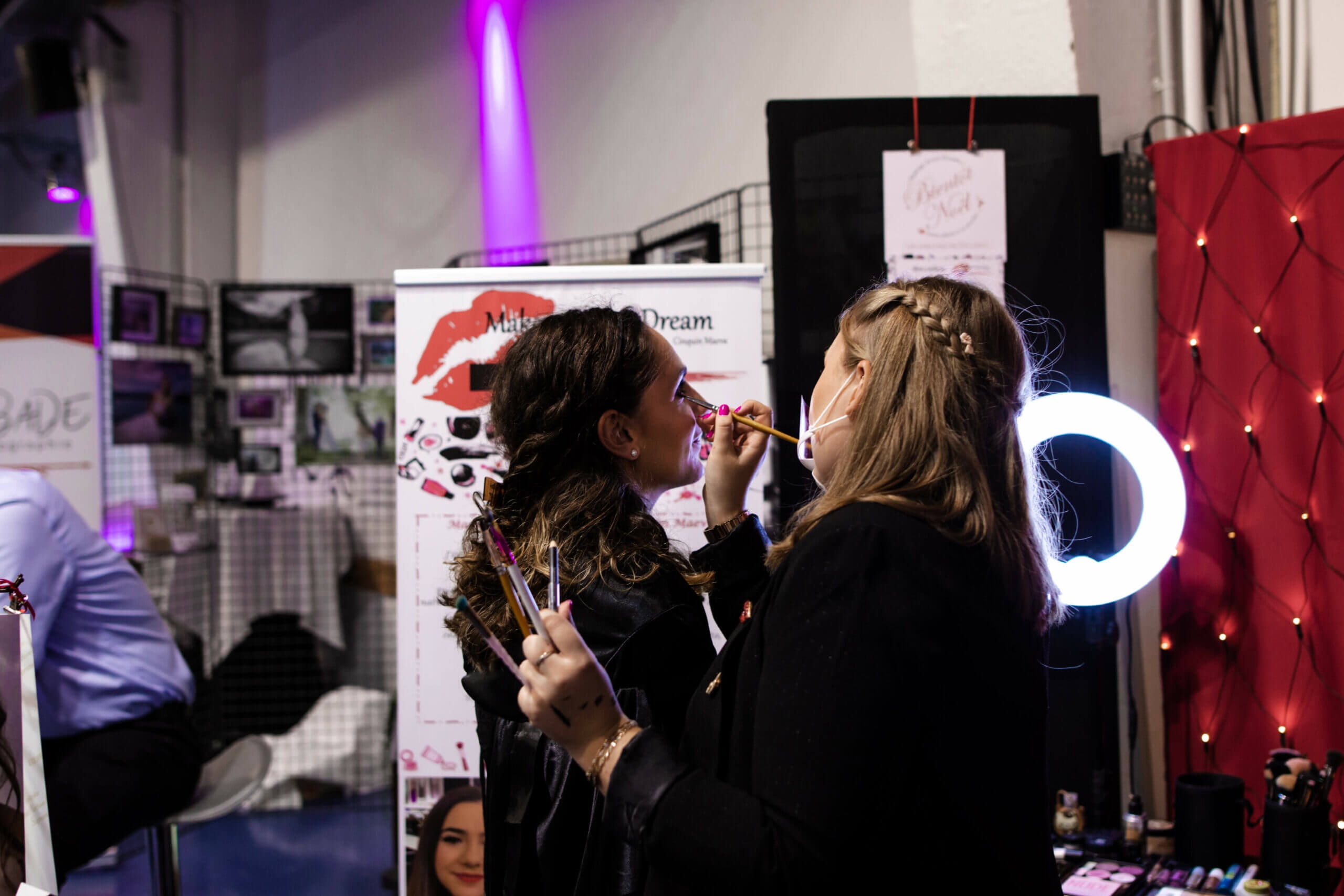 Deux femmes engagées dans l'application de maquillage dans un cadre de beauté animé, avec un fond décoratif rempli de matériaux promotionnels et de couleurs vives, créant une ambiance expressive et vibrante.
