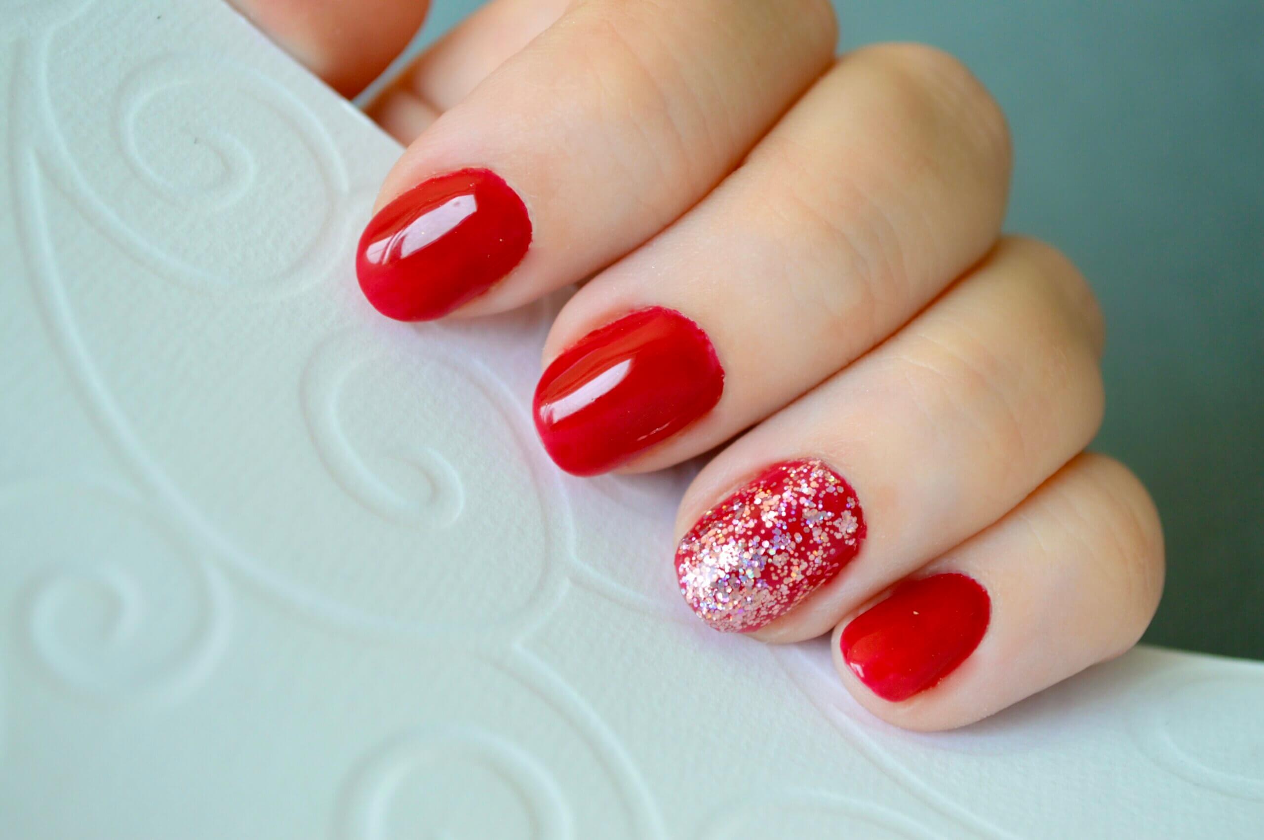 Gros plan d'une main avec une manucure élégante, des ongles laqués en rouge vif et un ongle décoré de paillettes argentées et roses, sur une surface blanche texturée.