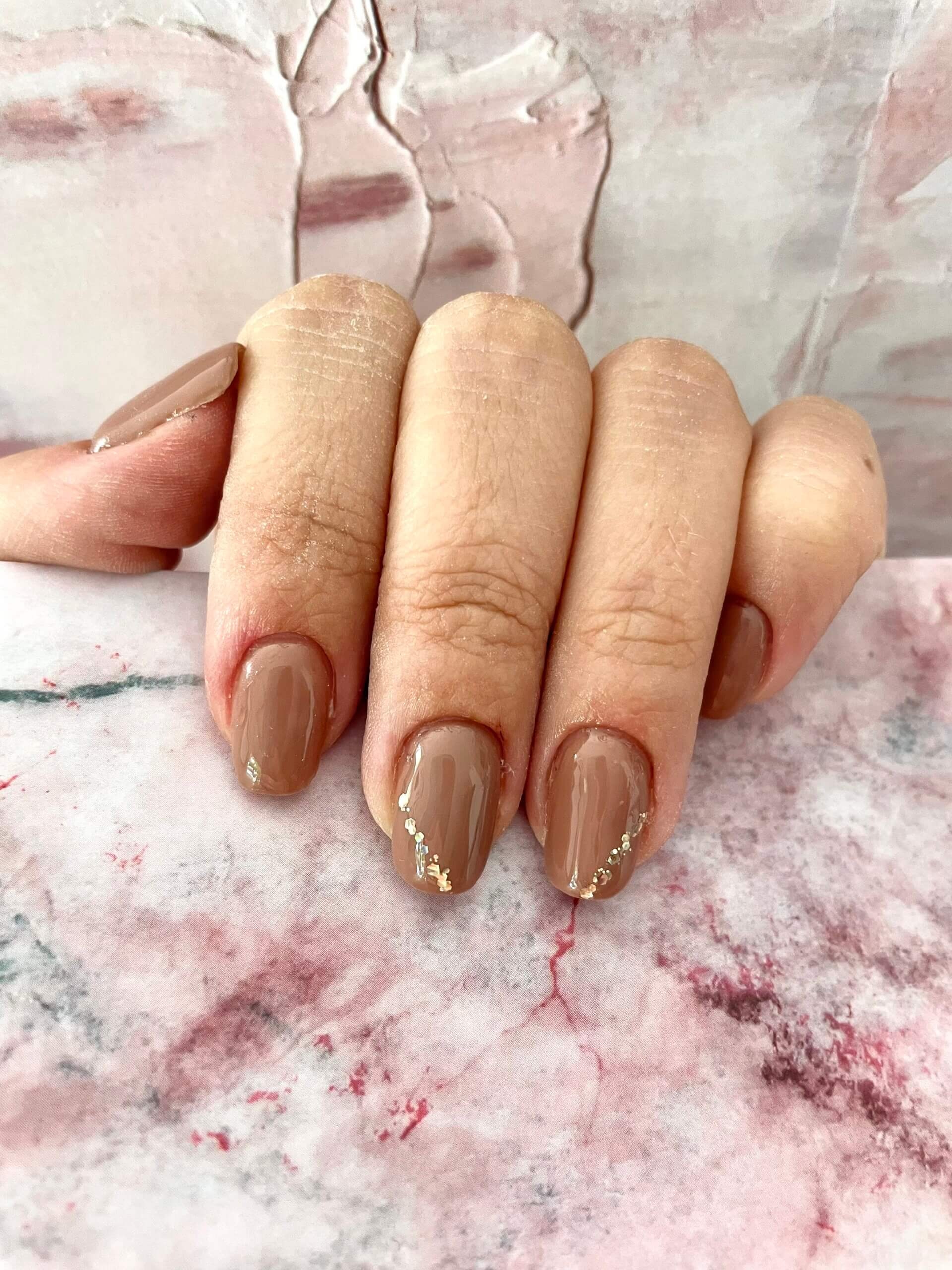 Gros plan d'une main avec des ongles parfaitement manucurés, peints dans une teinte beige neutre, avec des accents dorés. La main repose sur un fond texturé aux tons doux de rose et de blanc, mettant en valeur l'esthétique globale.