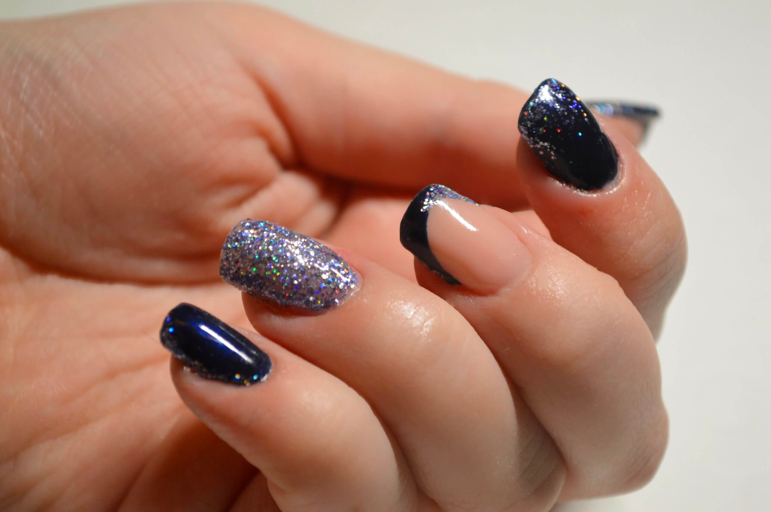 Gros plan d'une main montrant une manucure stylée avec des ongles bleu marine brillant, des paillettes étincelantes, et un ongle nude avec une pointe blanche délicate, le tout sur un fond clair.