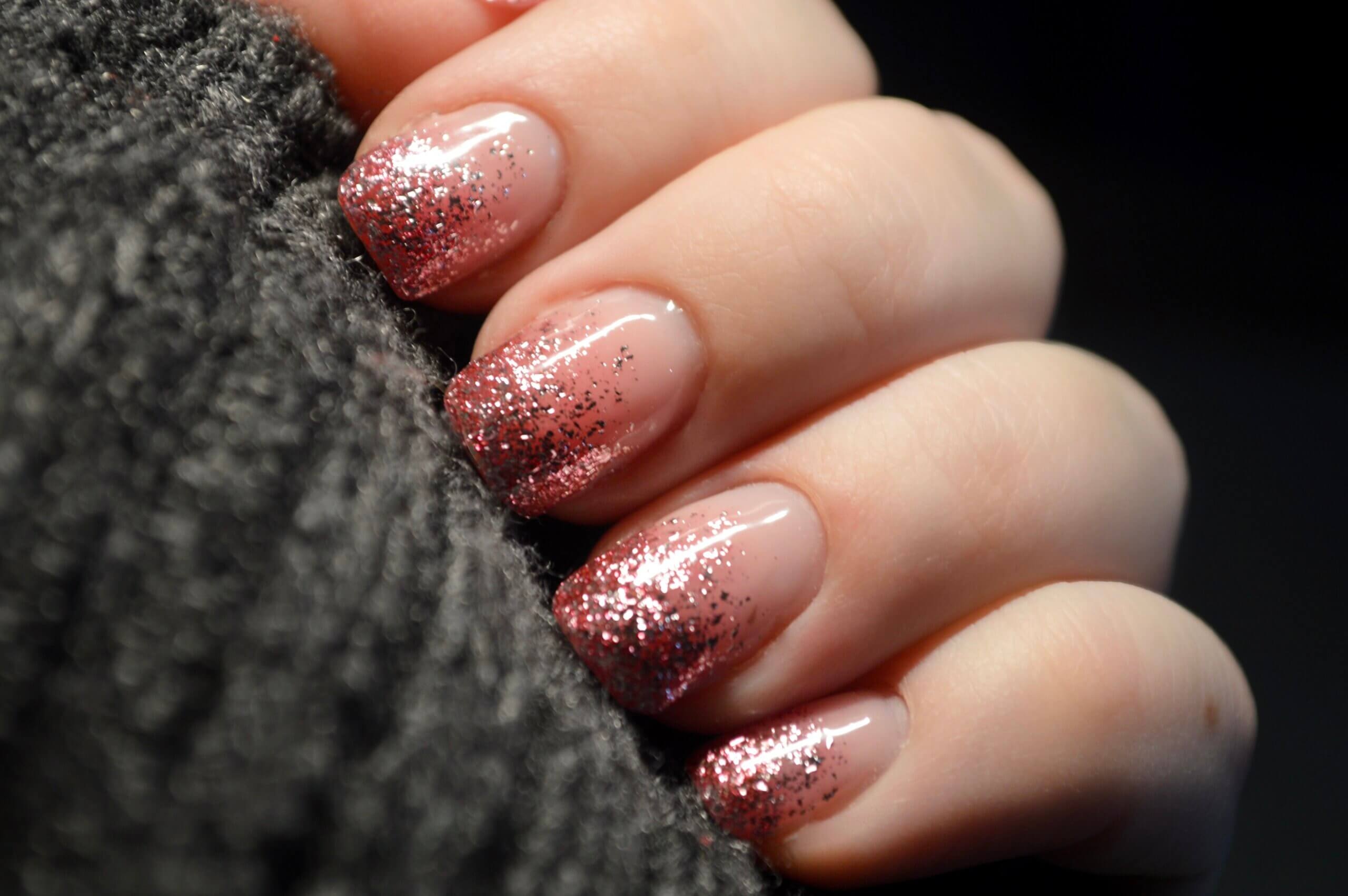 Gros plan sur une main affichant des ongles manucurés avec un design dégradé, une base nude brillante se transformant en rose pailleté aux pointes, reposant partiellement sur une surface texturée foncée.