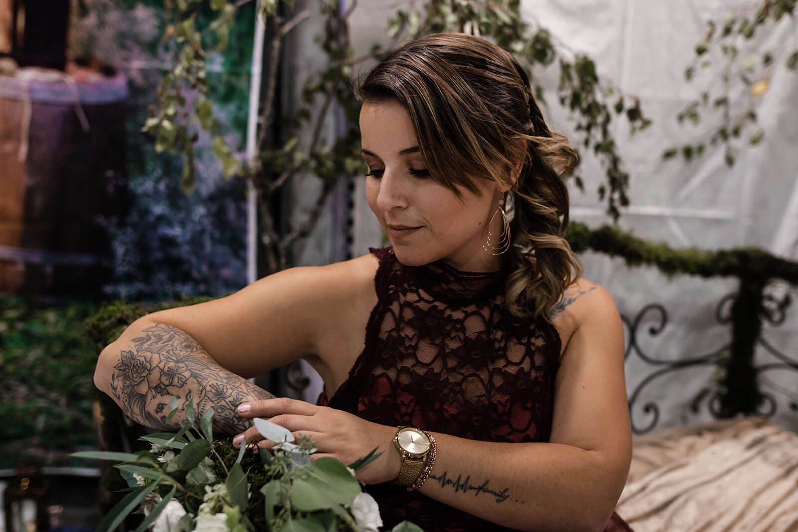 Une femme aux cheveux longs et ondulés, vêtue d'une robe en dentelle sombre, arrange doucement des fleurs dans un cadre naturel serein, révélant des tatouages artistiques sur ses bras.