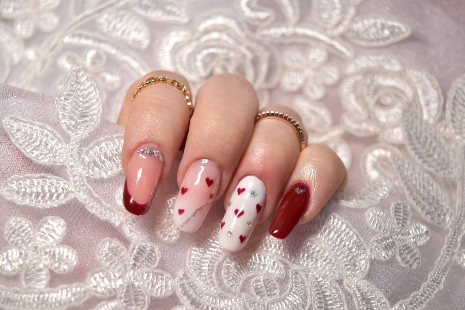 Gros plan d'une main bien manucurée avec des ongles ornés de nail art, des finitions rouge profond et une base rose clair décorée de petits cœurs rouges et de gemmes colorées, posée sur un fond délicat en dentelle blanche.