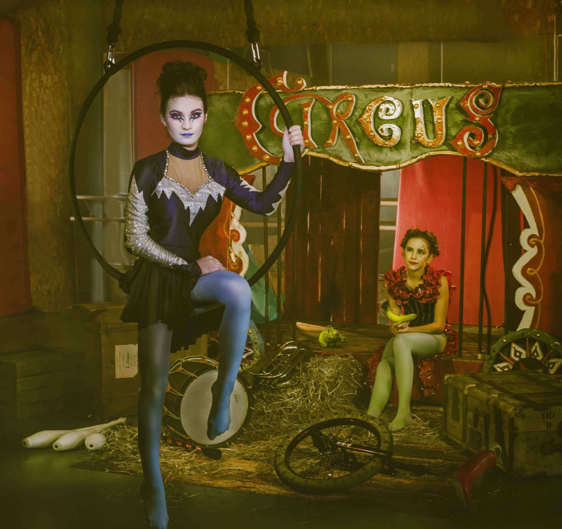 Une image d'un cirque avec deux artistes, l'un posant élégamment dans un cerceau avec un costume noir et argent, et l'autre assis sur une caisse en bois, vêtu d'une tenue rouge éclatante et tenant des bananes, le tout dans un décor coloré de cirque.