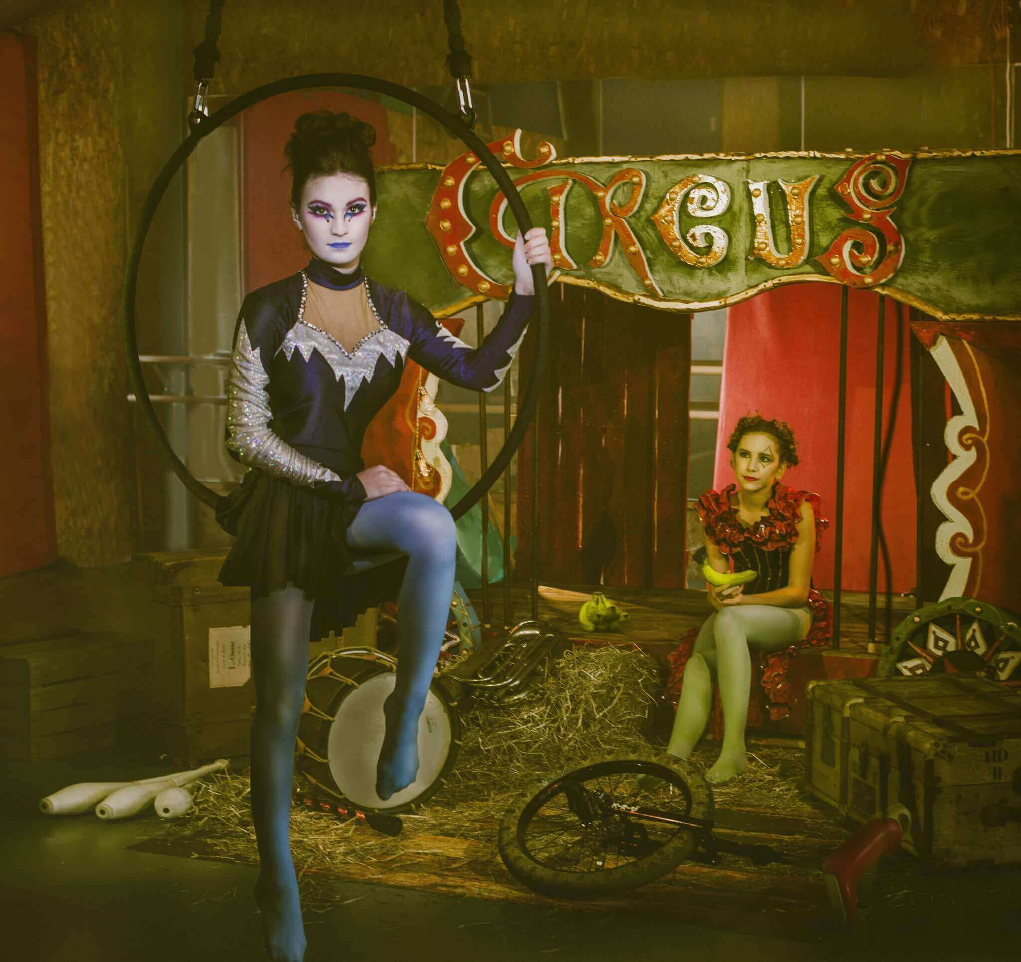 Une image d'un cirque avec deux artistes, l'un posant élégamment dans un cerceau avec un costume noir et argent, et l'autre assis sur une caisse en bois, vêtu d'une tenue rouge éclatante et tenant des bananes, le tout dans un décor coloré de cirque.