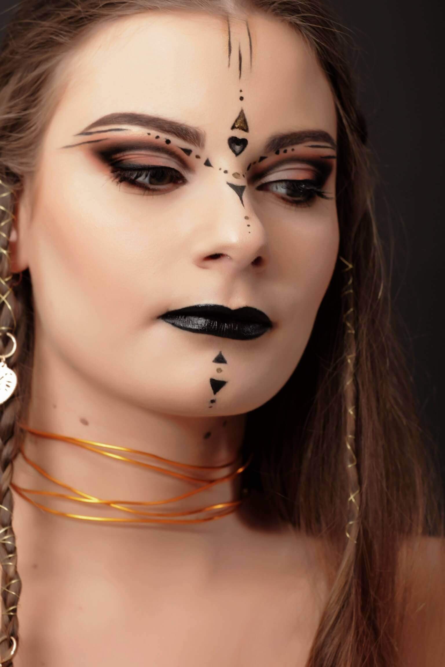 Un portrait en gros plan d'une femme avec un maquillage dramatique et des accessoires uniques, mettant en valeur des sourcils accentués et des designs d'eyeliner complexes, entourés d'une ombre brune chaude, des lèvres noires profondes, et des motifs géométriques frappants en noir et or sur son front et son menton.
