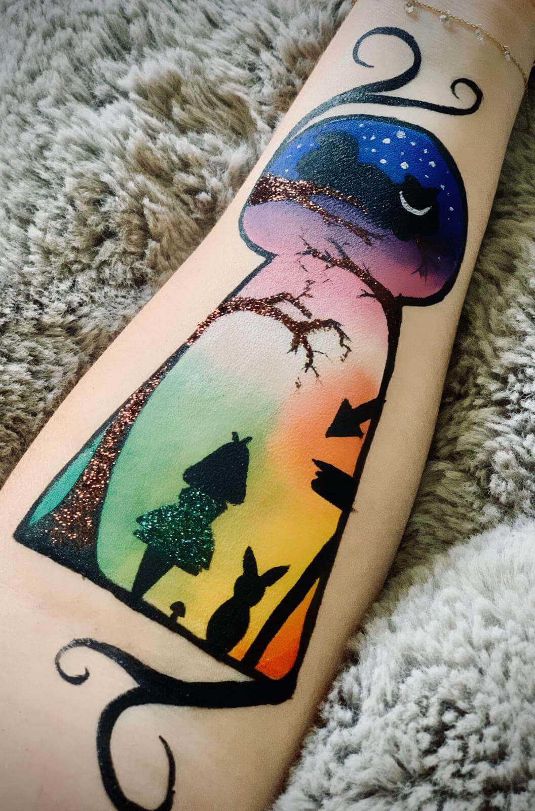 Tatouage coloré sur un avant-bras représentant une scène fantaisiste avec un grand champignon, un ciel nocturne étoilé, des branches d'arbres enchanteurs et des silhouettes de personnages, y compris un petit personnage et un lapin, sur un fond dégradé orange, rose et bleu.