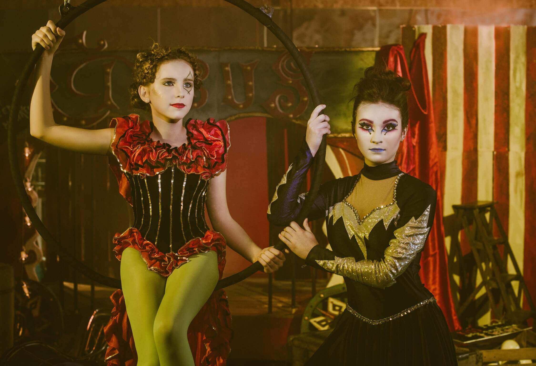 Deux artistes de cirque présentant des costumes vibrants dans un cadre de cirque animé. À gauche, une fille aux cheveux bouclés dans un costume de corset rouge et noir pose avec assurance sur un anneau. À droite, une fille dans une tenue élégante sombre tient l'anneau, ornée d'un maquillage audacieux et expressif sur fond de tissu rayé et de accessoires de cirque.