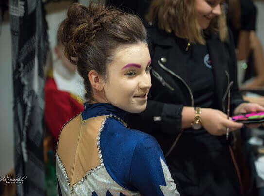 Une jeune artiste avec un chignon élégant, des sourcils violets audacieux et un costume en sequins colorés sourit en se retournant, dégageant de la confiance dans un cadre de performance vivant.