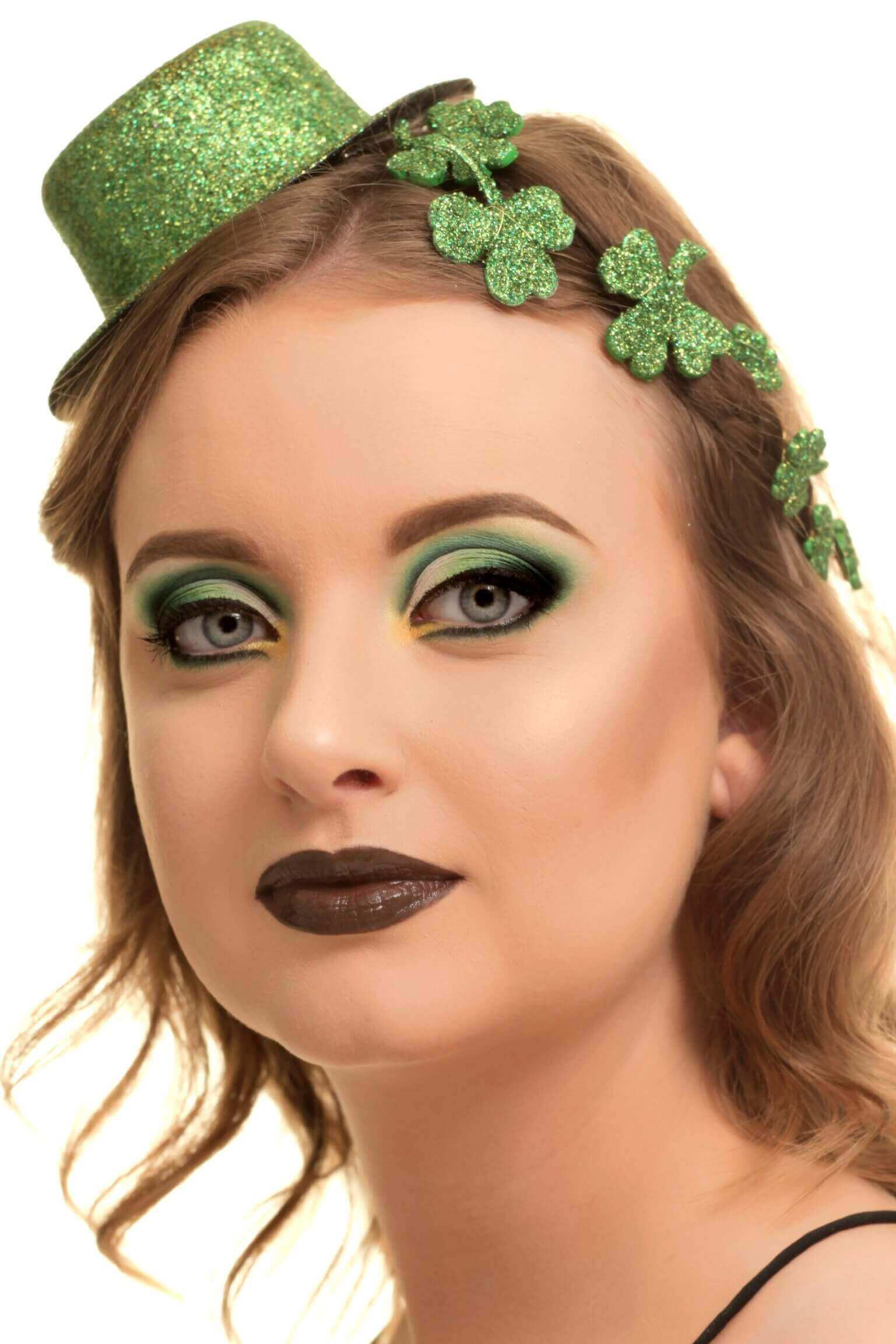 Portrait en gros plan d'une femme avec des fards à paupières verts et jaunes, des lèvres foncées et un mini haut-de-forme vert pailleté orné de trèfles, sur fond blanc.