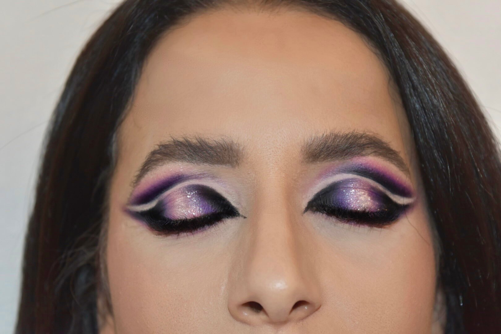 Gros plan d'yeux fermés présentant un design de maquillage complexe avec des formes géométriques en violet et noir, rehaussé de paillettes et d'un eye-liner détaillé.