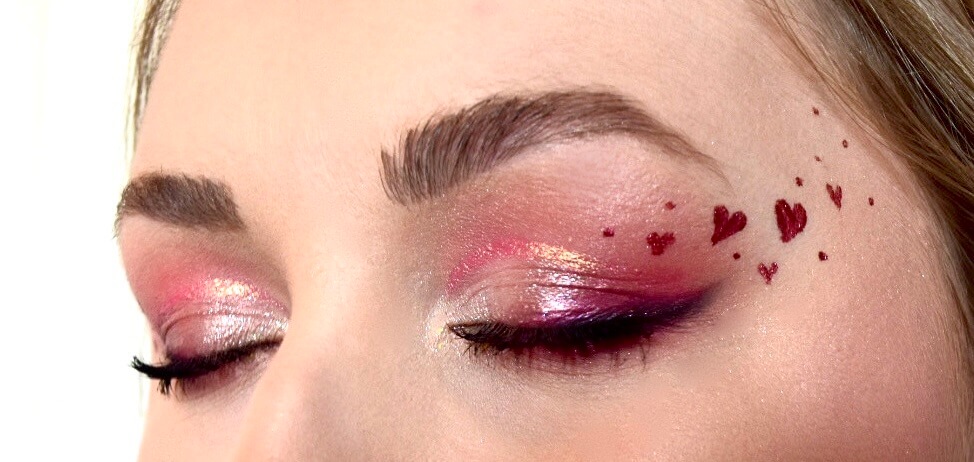 Gros plan sur un œil mettant en valeur un maquillage artistique avec des teintes douces de rose sur la paupière et des accents scintillants, ornés de petites formes de cœur rouges sur la paupière inférieure et des sourcils bien entretenus, soulignant un look de beauté ludique et détaillé.